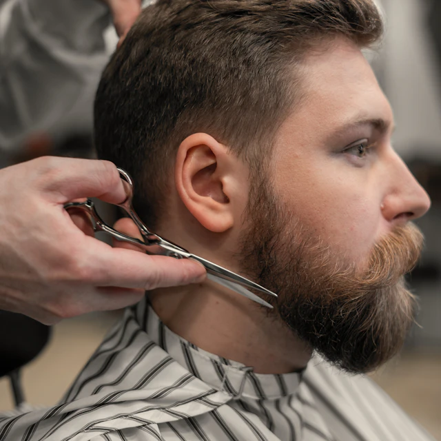 A barber trimming a man's beard with scissors in a salon