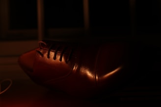 A close-up view of a brown leather shoe, with details of the laces and the polished surface highlighted by the dim lighting. The background is dark, enhancing the sheen on the shoe.