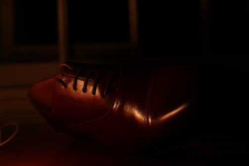 A close-up view of a brown leather shoe, with details of the laces and the polished surface highlighted by the dim lighting. The background is dark, enhancing the sheen on the shoe.