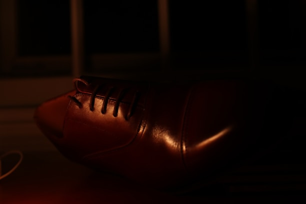 A close-up view of a brown leather shoe, with details of the laces and the polished surface highlighted by the dim lighting. The background is dark, enhancing the sheen on the shoe.
