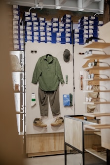 A retail display inside a store featuring outdoor or casual clothing and accessories arranged on a white board. Items include a green long-sleeve shirt, gray pants, a pair of green and beige shoes, a green hat, sunglasses, a pair of white socks, a small green item, and a blue pouch. In the background, shelves are stacked with blue and white shoe boxes.