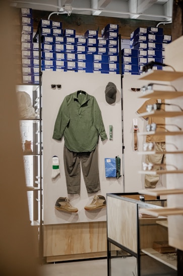 A retail display inside a store featuring outdoor or casual clothing and accessories arranged on a white board. Items include a green long-sleeve shirt, gray pants, a pair of green and beige shoes, a green hat, sunglasses, a pair of white socks, a small green item, and a blue pouch. In the background, shelves are stacked with blue and white shoe boxes.