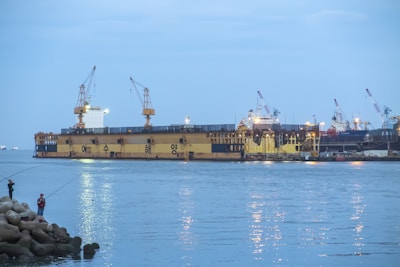 Large industrial ship docked near the shore with multiple cranes on board, surrounded by calm water. Two people are fishing from the rocky shore in the foreground. The sky is overcast, contributing to a subdued atmosphere.
