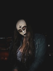 A person wearing a mask with a cross and blackened eye designs, long hair, and a plaid outfit stands in a dark environment. The lighting is low, emphasizing the eerie features of the mask.