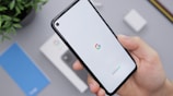A hand holding a smartphone with a white screen displaying the Google logo and the word 'Android'. In the background, there's a smartphone box, a charger, a blue booklet, and a plant, all situated on a light gray surface.