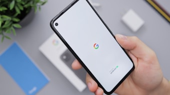 A hand holding a smartphone with a white screen displaying the Google logo and the word 'Android'. In the background, there's a smartphone box, a charger, a blue booklet, and a plant, all situated on a light gray surface.