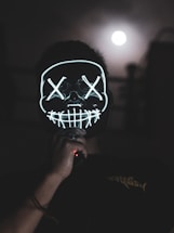 A person is wearing a glowing mask with stitched mouth and cross symbols over the eyes, standing in a dark setting. The mask is illuminated with neon lights. In the background, the full moon appears in a cloudy night sky, creating an eerie atmosphere.