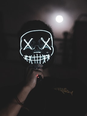 A person is wearing a glowing mask with stitched mouth and cross symbols over the eyes, standing in a dark setting. The mask is illuminated with neon lights. In the background, the full moon appears in a cloudy night sky, creating an eerie atmosphere.