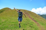 A person is joyfully running on a grassy hill under a clear blue sky. The path on the hill is slightly worn, indicating frequent use. The scene conveys a sense of freedom and adventure, with the hiker wearing casual outdoor attire and a backpack.