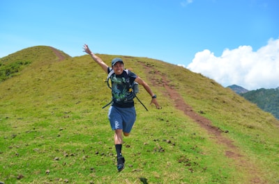 A person is joyfully running on a grassy hill under a clear blue sky. The path on the hill is slightly worn, indicating frequent use. The scene conveys a sense of freedom and adventure, with the hiker wearing casual outdoor attire and a backpack.