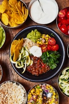 A bowl filled with a variety of colorful ingredients including guacamole, cherry tomatoes, curly zucchini, a dollop of white sauce, kale, corn mixed with red onions, and a hearty bean chili. Surrounding the bowl are several small dishes containing tortilla chips, white sauce, cherry tomatoes, cooked rice, and corn salad. The scene is set on a rustic wooden surface.