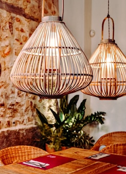Two large, decorative woven pendant lights hang above a wooden dining table. The lights emit a warm glow, creating a cozy and inviting atmosphere. In the background, there is a stone wall with visible textures, and a few potted plants are placed near the wall. The table is set with red placemats and neatly folded napkins.
