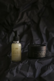 A bottle of face serum with a dropper lid and a jar of day cream are placed on a dark, crinkled fabric background. The bottle is a frosted glass with a black dropper, while the jar is dark and possibly metallic or plastic with elegant labels. The lighting is soft, creating a moody and sophisticated atmosphere.