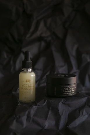 A bottle of face serum with a dropper lid and a jar of day cream are placed on a dark, crinkled fabric background. The bottle is a frosted glass with a black dropper, while the jar is dark and possibly metallic or plastic with elegant labels. The lighting is soft, creating a moody and sophisticated atmosphere.