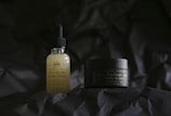 Two skincare products are positioned on a dark, textured background. The bottle on the left is a face serum with a dropper lid, featuring a label that includes the words 'Face Serum' and 'CBD'. On the right, a small jar labeled 'Day Cream' accompanies it, providing a sense of a skincare routine.