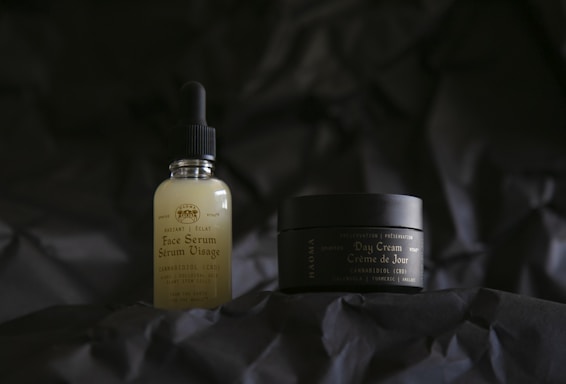 Two skincare products are positioned on a dark, textured background. The bottle on the left is a face serum with a dropper lid, featuring a label that includes the words 'Face Serum' and 'CBD'. On the right, a small jar labeled 'Day Cream' accompanies it, providing a sense of a skincare routine.