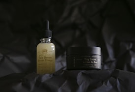 Two skincare products are positioned on a dark, textured background. The bottle on the left is a face serum with a dropper lid, featuring a label that includes the words 'Face Serum' and 'CBD'. On the right, a small jar labeled 'Day Cream' accompanies it, providing a sense of a skincare routine.