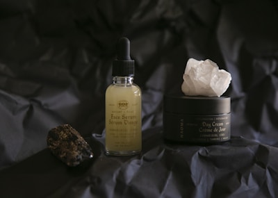 A bottle of face serum with a dropper and a jar of day cream are displayed on a dark, crumpled surface. The serum bottle is transparent with a light yellow liquid inside, while the day cream jar is black with a large white crystal on top. A dark, textured stone is positioned to the left.