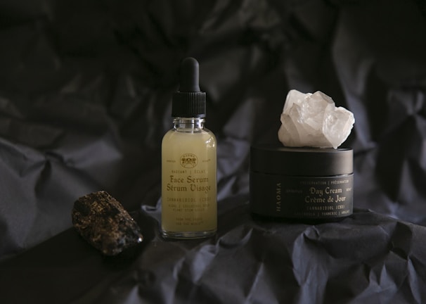 A bottle of face serum with a dropper and a jar of day cream are displayed on a dark, crumpled surface. The serum bottle is transparent with a light yellow liquid inside, while the day cream jar is black with a large white crystal on top. A dark, textured stone is positioned to the left.
