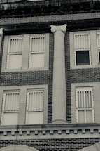 A brick building facade with two rows of rectangular windows featuring vertical blinds. A prominent classical column with a decorative capital is positioned between the windows.