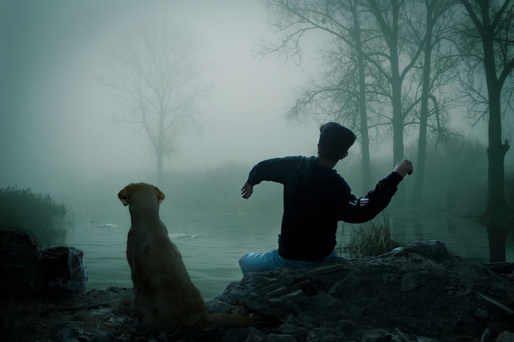 man in black jacket and blue denim jeans sitting on ground beside brown short coated dog