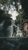 A person stands in shallow water facing a waterfall, surrounded by dense greenery and trees. The individual is wearing a backpack and holding a helmet, suggesting an adventurous or exploring activity. The waterfall cascades down rocks with a misty effect, creating a refreshing natural scene.