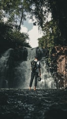 A person stands in shallow water facing a waterfall, surrounded by dense greenery and trees. The individual is wearing a backpack and holding a helmet, suggesting an adventurous or exploring activity. The waterfall cascades down rocks with a misty effect, creating a refreshing natural scene.