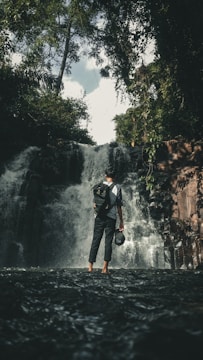 A person stands in shallow water facing a waterfall, surrounded by dense greenery and trees. The individual is wearing a backpack and holding a helmet, suggesting an adventurous or exploring activity. The waterfall cascades down rocks with a misty effect, creating a refreshing natural scene.