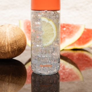 A transparent water bottle with an orange cap is filled with ice and a lemon slice inside. It is positioned on a reflective surface with a coconut and watermelon slices in the background.