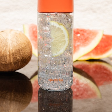 A transparent water bottle with an orange cap is filled with ice and a lemon slice inside. It is positioned on a reflective surface with a coconut and watermelon slices in the background.