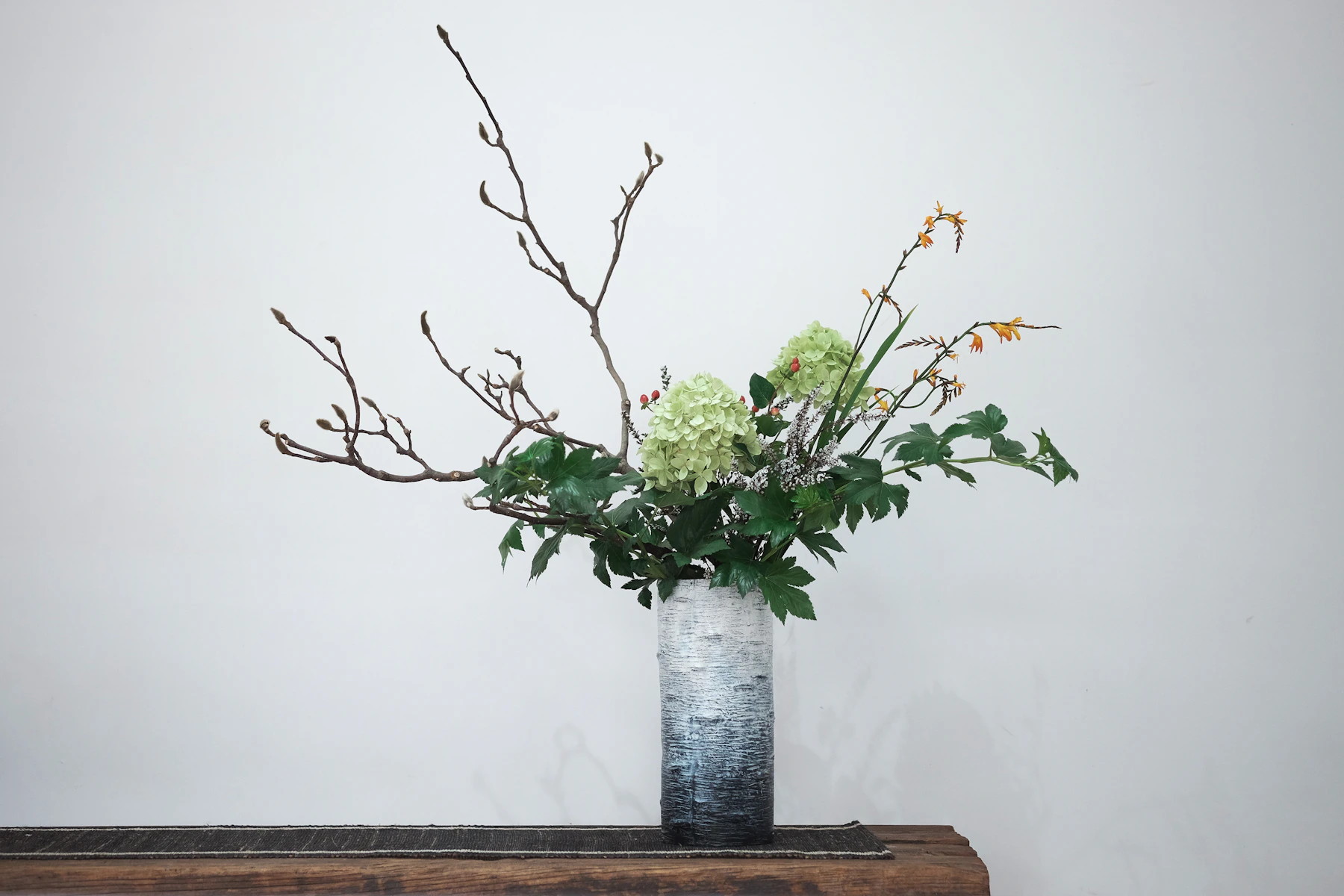A budding bare branch, green hydrangeas and leaves arranged in a tall gray cylinder vase on a wooden bench