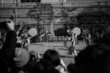 A black-and-white photograph features a group of traditional dancers performing in an outdoor setting. The dancers wear intricate costumes and play large drums, while the audience watches the performance. Tall trees and a stone wall backdrop enhance the cultural ambiance.