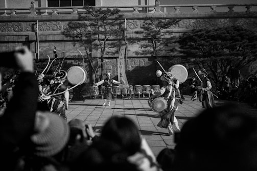A black-and-white photograph features a group of traditional dancers performing in an outdoor setting. The dancers wear intricate costumes and play large drums, while the audience watches the performance. Tall trees and a stone wall backdrop enhance the cultural ambiance.