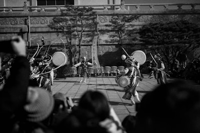 A black-and-white photograph features a group of traditional dancers performing in an outdoor setting. The dancers wear intricate costumes and play large drums, while the audience watches the performance. Tall trees and a stone wall backdrop enhance the cultural ambiance.