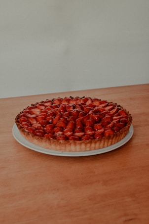 A beautifully baked tart topped with glistening strawberries arranged neatly on a white plate. The tart has a golden-brown crust and is set on a wooden surface against a plain background.
