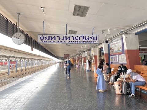 A spacious train station platform with a prominent sign in both Thai and English. People are sitting on benches and standing around, some wearing masks. The platform extends into the distance with tracks visible on the left, and there are various posters and signs along the walls.