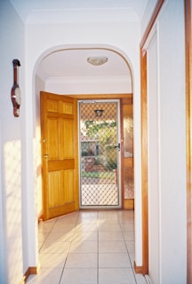 A hallway featuring a tiled floor leading to an open wooden door with a metal mesh security screen. Natural light casts shadows on the floor and walls, creating a warm atmosphere. Wall accents include a hanging clock.