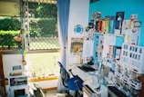 A colorful and vibrant home office with a desk cluttered with books, toys, and stationery. A bright blue accent wall features shelves holding an array of books organized by color, along with decorative items like a globe and a motivational sign. A window with a curtain looks out to a lush green garden, allowing natural light to filter in.