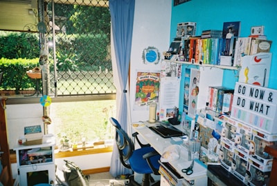 A colorful and vibrant home office with a desk cluttered with books, toys, and stationery. A bright blue accent wall features shelves holding an array of books organized by color, along with decorative items like a globe and a motivational sign. A window with a curtain looks out to a lush green garden, allowing natural light to filter in.
