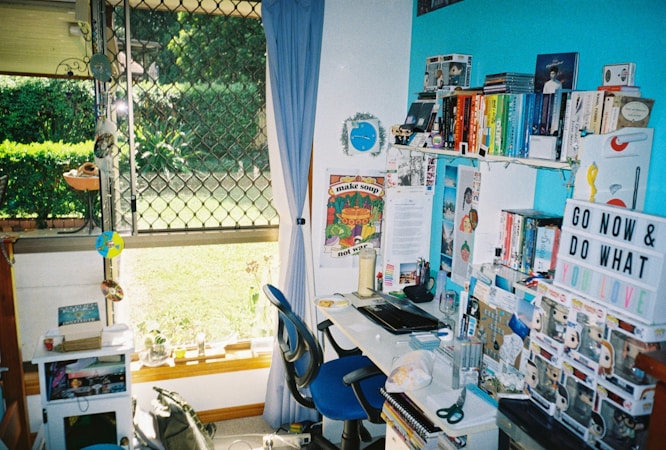 A colorful and vibrant home office with a desk cluttered with books, toys, and stationery. A bright blue accent wall features shelves holding an array of books organized by color, along with decorative items like a globe and a motivational sign. A window with a curtain looks out to a lush green garden, allowing natural light to filter in.