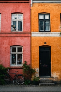 Side-by-side brick buildings feature contrasting colors, with one in a muted red and the other in a vibrant orange. Each facade has white-framed windows and finely detailed molding beneath the roof. A black bicycle rests next to the red building, and potted plants adorn the area near both structures, adding a touch of greenery.