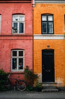 Side-by-side brick buildings feature contrasting colors, with one in a muted red and the other in a vibrant orange. Each facade has white-framed windows and finely detailed molding beneath the roof. A black bicycle rests next to the red building, and potted plants adorn the area near both structures, adding a touch of greenery.