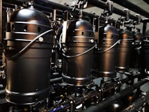 A row of industrial black stage lights is suspended from a metal framework. The lights feature cylindrical housings with handles and ventilation holes, suggesting they are meant for a theatrical or concert setting. The metallic surface and compact design indicate a robust construction.