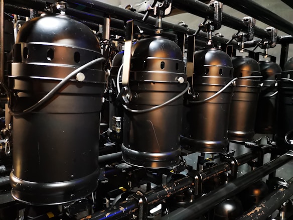 A row of industrial black stage lights is suspended from a metal framework. The lights feature cylindrical housings with handles and ventilation holes, suggesting they are meant for a theatrical or concert setting. The metallic surface and compact design indicate a robust construction.