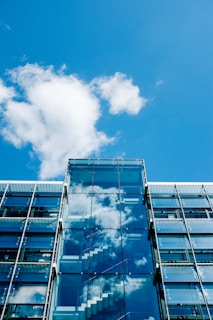 A modern glass building with reflections of white clouds and a blue sky on its surface. The facade prominently features transparent glass panels and visible internal staircases.