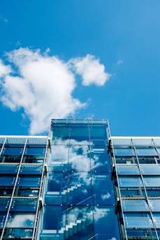 A modern glass building with reflections of white clouds and a blue sky on its surface. The facade prominently features transparent glass panels and visible internal staircases.