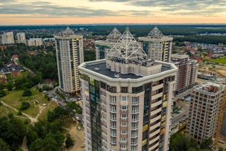 High-rise residential buildings with unique architectural designs feature prominently, surrounded by greenery and an urban landscape under a cloudy sky.