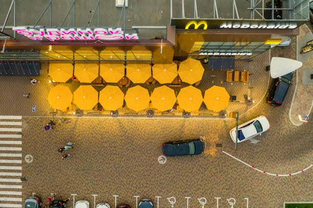 An aerial view shows a patio with several yellow umbrellas arranged in a neat grid pattern. The outdoor seating area belongs to a fast-food restaurant, as indicated by the recognizable branding. Surrounding the patio are parked cars and pedestrians walking on a brick-patterned pavement. The scene is colorful and vibrant, with the bold yellow of the umbrellas contrasting with the darker shades of the pavement and vehicles.