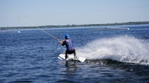 A person wearing a blue helmet and life jacket is wakeboarding on a large body of water. They are creating a splash as they move, holding onto a rope connected to a pulley system above. In the background, there are floating structures and the horizon is visible.