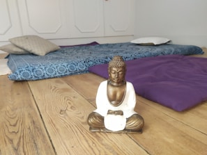 A serene room with a wooden floor, featuring a small statue of a Buddha sitting in a meditative pose. Behind the statue, there are cushions and a blue patterned mat arranged on the floor, creating a peaceful and relaxing atmosphere.